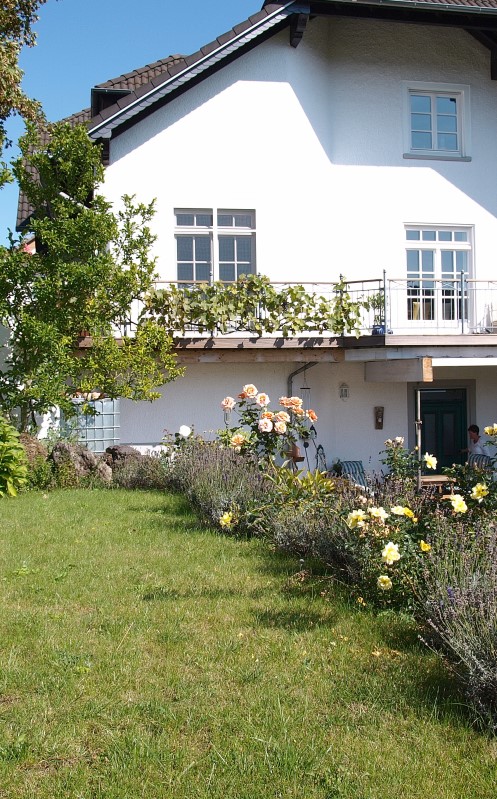 Ferienwohnung Zauberhaft Fam. Münch in Schalkenmehren - Außenansicht mit grüner Wiese, Garten und Rosenbüschen sowie Terrasse und Balkon